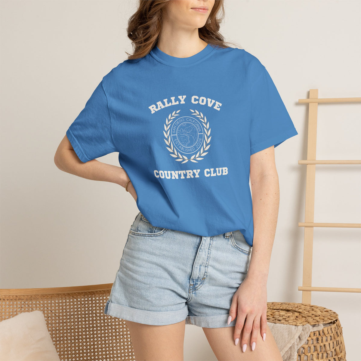 A woman wearing a blue short-sleeve graphic t-shirt with a white 'Rally Cove Country Club' emblem on the chest, paired with light-wash denim shorts in a brightly lit indoor setting.