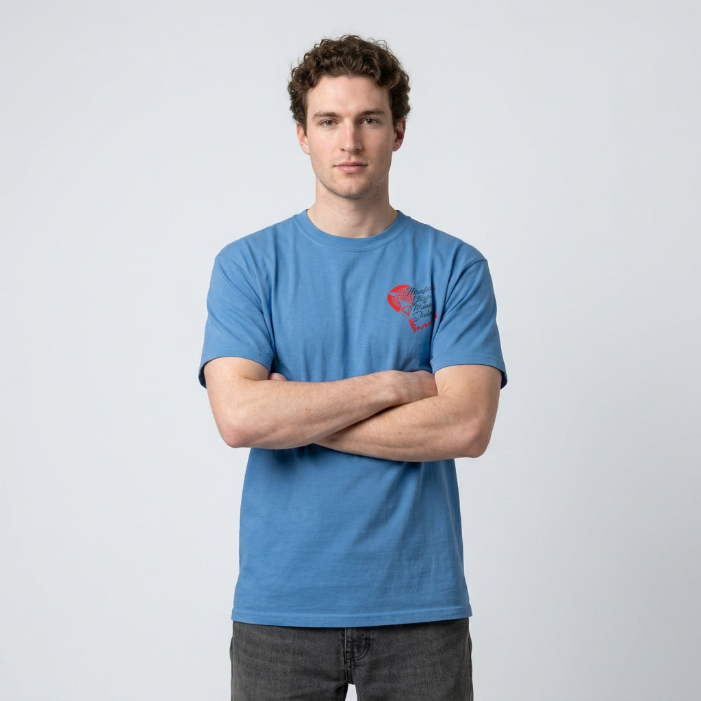 A man wearing a blue crewneck t-shirt with a small red padel graphic on the left chest, standing with arms crossed against a light gray background.