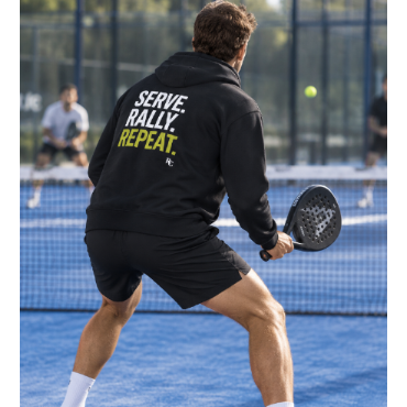 A man on a blue tennis court wearing a black hoodie with 'SERVE, RALLY, REPEAT.' printed in white and yellow on the back, black athletic shorts, white socks, and white sneakers, holding a paddle racket.
